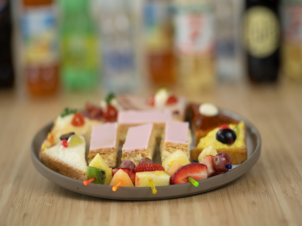 Im Vordergrund ist ein Teller mit belegten Broten, Früchtespiessen und Cremeschnitten, im Hintergrund verschiedene Softgetränke auf einem Holztisch.