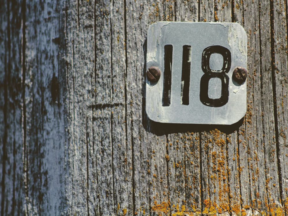 Ein Metallschildchen ist mit 118 beschriftet und mit zwei rostigen Nägeln an einer Holzwand befestigt.